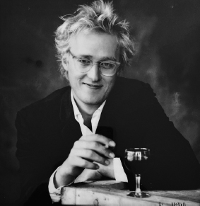Start of image description, A promotional image captures Adrian in his early twenties with a shock of blond hair. He is smiling and reaching for a glass of red wine. He wears an open-necked white shirt, black jacket, and a pair of plastic-frame glasses., end of image description