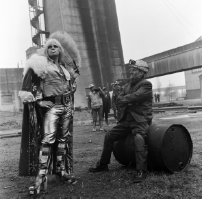 Start of image description, The flamboyant wrestler Adrian Street poses with his father at the Blaina colliery in November 1974. Adrian has long blond hair, face paint and sequins, an open-necked shirt, a feather boa trimmed cape, shiny silver trousers, and high-heeled boots. He holds up a feather fan. His father sits on an oil barrel wearing a mining helmet, his smiling face smeared with coal dust., end of image description