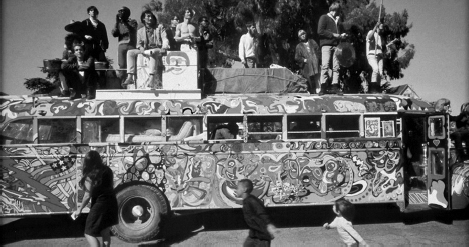 Start of image description, The colourful Merry Pranksters’ bus from the 1960s. The old school bus is parked up and various counter-culture characters are sitting and standing on the roof as children play on the ground., end of image description