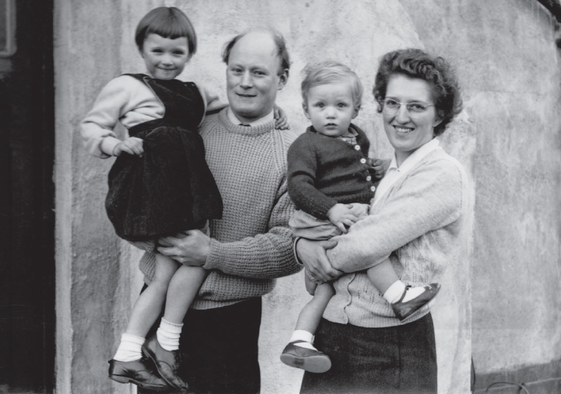 Start of image description, A photograph from 1958 shows baby Adrian in the arms of his mother, Dorothy. His father Fred holds up his 4 year old sister, Hilary. Adrian’s hair is styled with a side parting, Hilary’s hair is cut with a bowl style fringe, his mum has short, thick, wavy hair, and his dad is bald with hair on the sides., end of image description