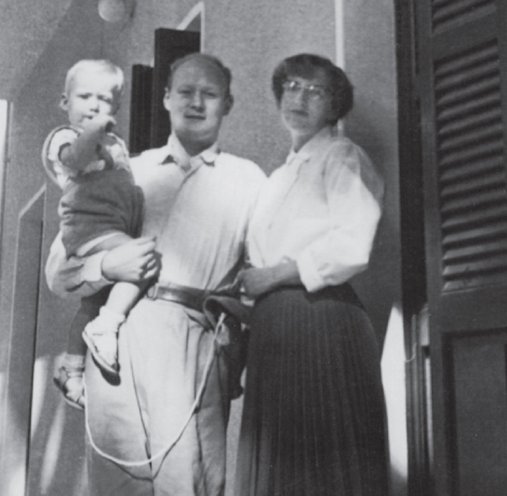 Start of image description, A young Adrian is held by his father as his mother stands beside them in Nicosia. Fred has a holstered gun at his hip and a lanyard is attached the gun., end of image description