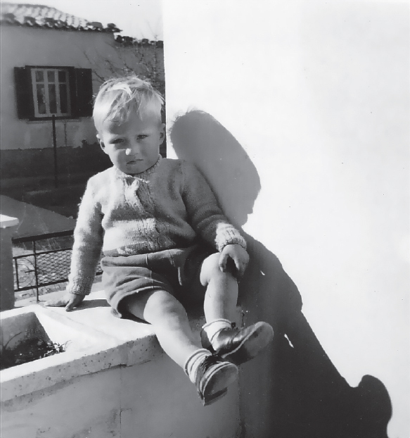 Start of image description, Adrian, aged around 2 years old, sits on a wall outside their home at the Wolseley Barracks in Nicosia. Adrian is frowning at the camera and wearing a sweater and a pair of shorts., end of image description