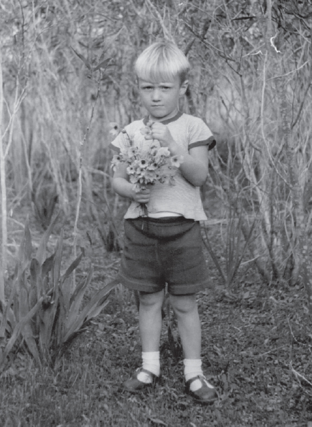 Start of image description, Adrian, aged around 3 years old, stands in a field holding a small bouquet of wildflowers. He has very light, blond hair and he is wearing a white t-shirt, dark shorts, and sandals with white socks., end of image description