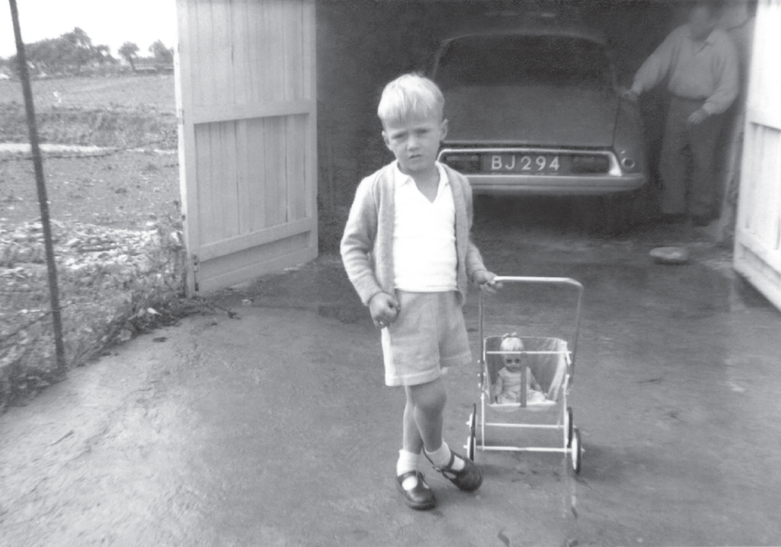 Start of image description, Adrian, aged around 4 years old, stands on the driveway outside the garage and pulls a pram with a baby doll inside. His father stands inside the garage, polished his beloved Citroen car., end of image description