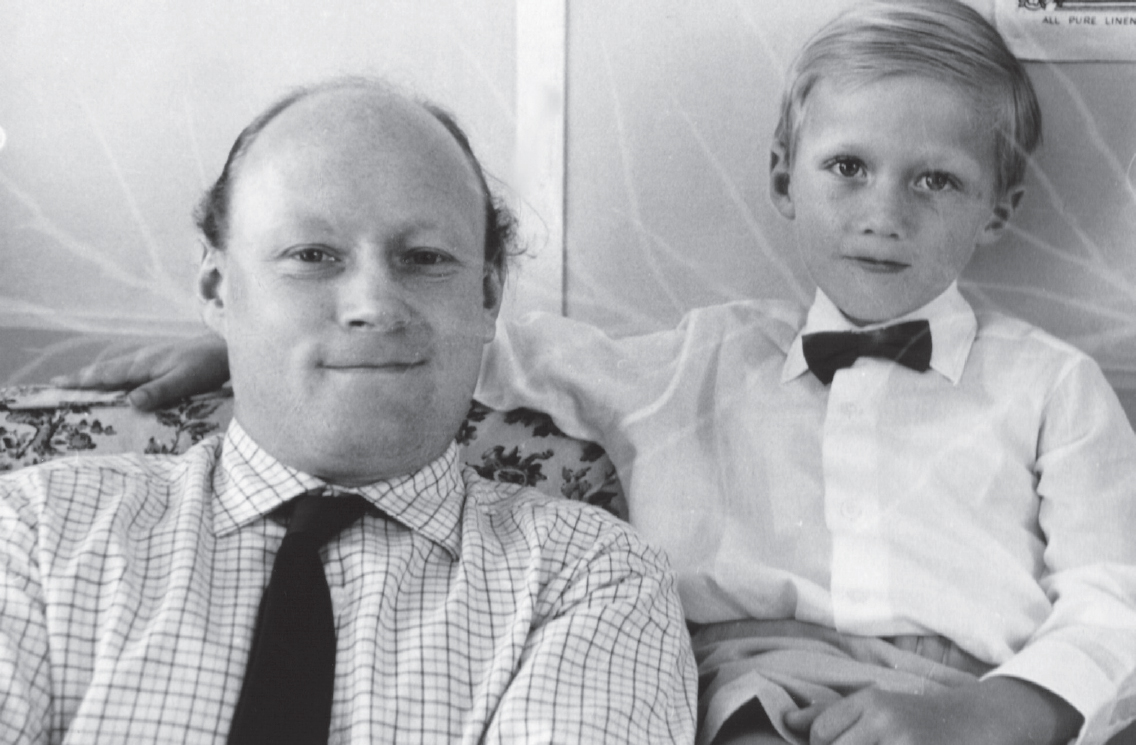 Start of image description, Adrian poses with his dad on the sofa of their home before the Christening of his young brother, Matthew. Adrian wears a white shirt with a black bowtie and rests his arm on the back of the sofa. His father wears a shirt and tie., end of image description