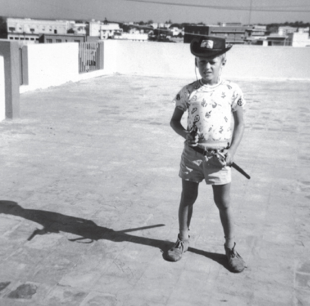 Start of image description, A young Adrian, aged around 8, poses on the roof of the family flat in Bahrain on a bright, sunny day. He is wearing a cowboy hat and pointing a gun. He has a sword in a scabbard at his hip., end of image description