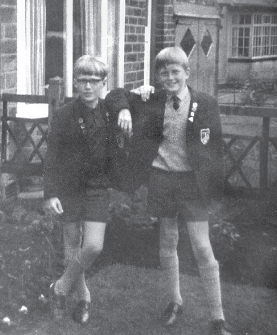 Start of image description, Adrian is pictured with a friend in their uniforms for Hutton Junior High School in Bradford in 1968, aged 11 years old. They are standing in the garden and Adrian, wearing glasses, leans on his friend’s shoulder. Both are wearing knee-high socks and mid-thigh-length shorts., end of image description