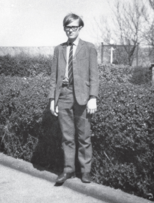 Start of image description, Adrian stands on the edge of a hedge at Pocklington Grammar School, aged around 16 years old. His hair is swept from left to right in a side parting and he wears thick, black framed glasses. He wears a suit and tie and poses with his arms by his side., end of image description