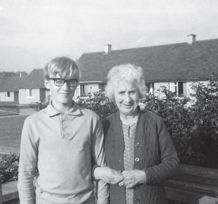 Start of image description, A teenage Adrian stands arm in arm with his grandmother outside her house. She has thick, white hair and wears a cardigan, and she is smiling. Adrian wears thick, black framed glasses and squints in the sunshine., end of image description