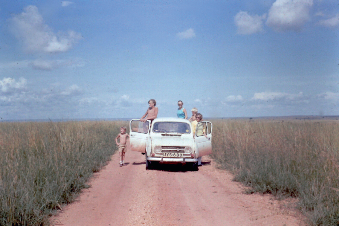 Start of image description, The Edmondson family stop during a safari in Kenya. The small Renault 4 car has its doors open and five figures are clustered around it, standing on the dusty road and looking out across the vast savannah., end of image description