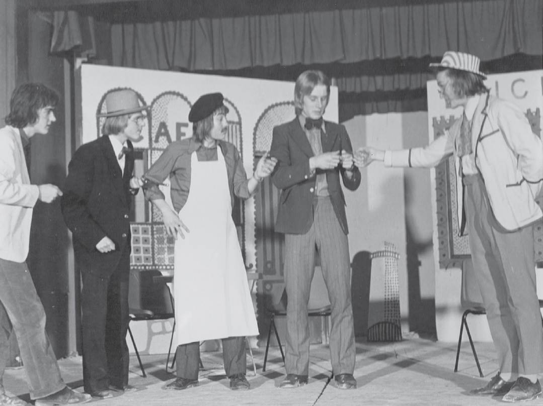 Start of image description, A performance of the play Rhinoceros at Pocklington School features 5 students on stage. Adrian is standing on the right, dressed in a light-coloured jacket, cravat, and summer hat. He is turned towards his co-stars and gesturing to them., end of image description