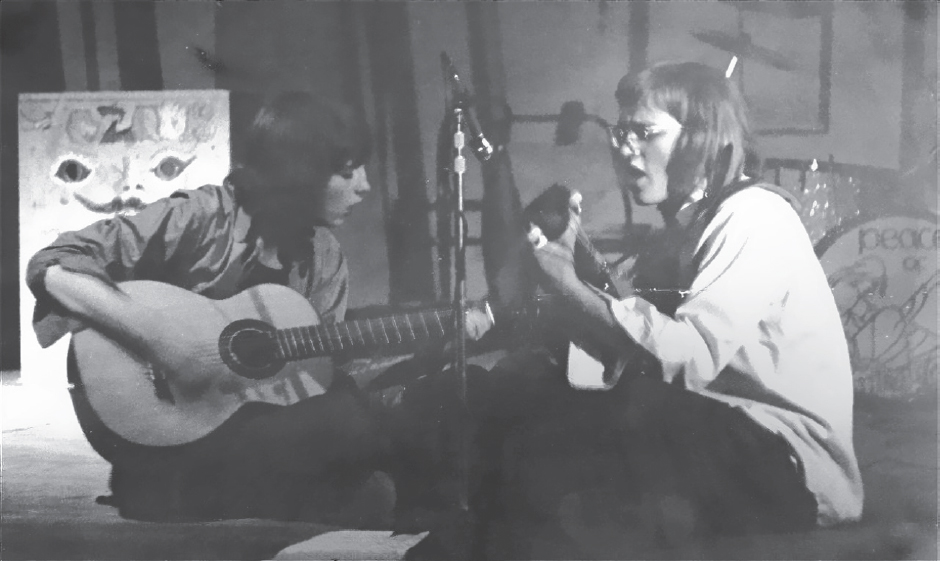 Start of image description, Adrian and his band-mate, Ian, sit and play guitar and sing during a rehearsal of their band, Peace of Thorn at Pocklington in 1975. On the right of the image in the background is a large box with a face painted on it., end of image description