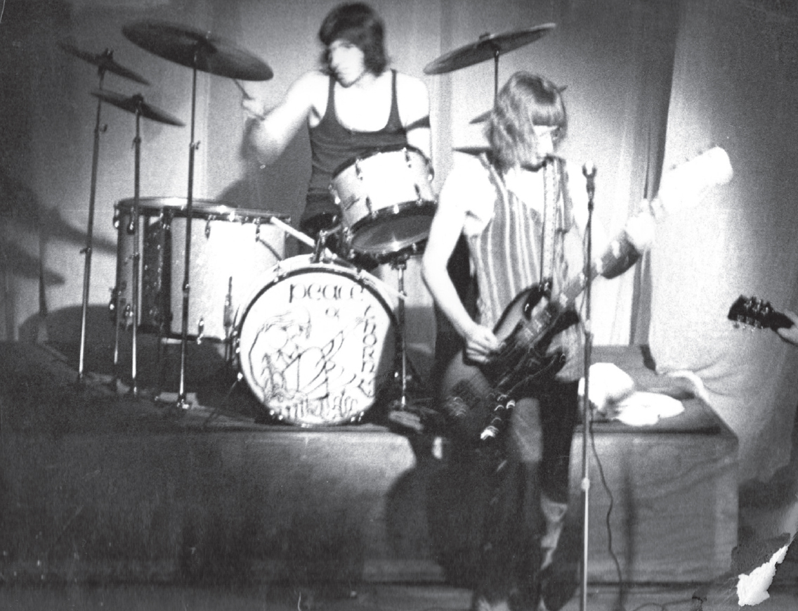 Start of image description, Adrian stands centre-stage and plays his bass guitar, his long hair framing his face. Ian plays the drums in the background. Another hand, holding the neck of a guitar, can be seen on the right of the photo., end of image description