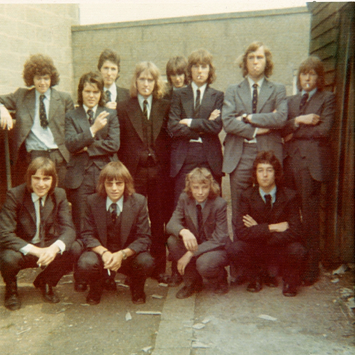 Start of image description, A group of 12 Pocklington students stand behind the bike shed and smoke. Adrian, wearing a dark three-piece suit, stands in the centre with shoulder length hair and his hands thrust in his pockets. A cigarette hangs from his lips., end of image description