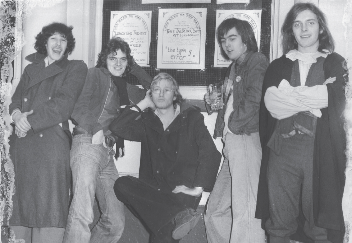 Start of image description, Adrian poses with his friends from Twentieth Century Coyote at The Band on the Wall in Manchester in 1976. Adrian sits in the centre with Lloyd Peters and Mark Dewison to his right and Mike Readfern and Rik Mayall to his left. Mayall has shoulder length hair and wears a long cape., end of image description