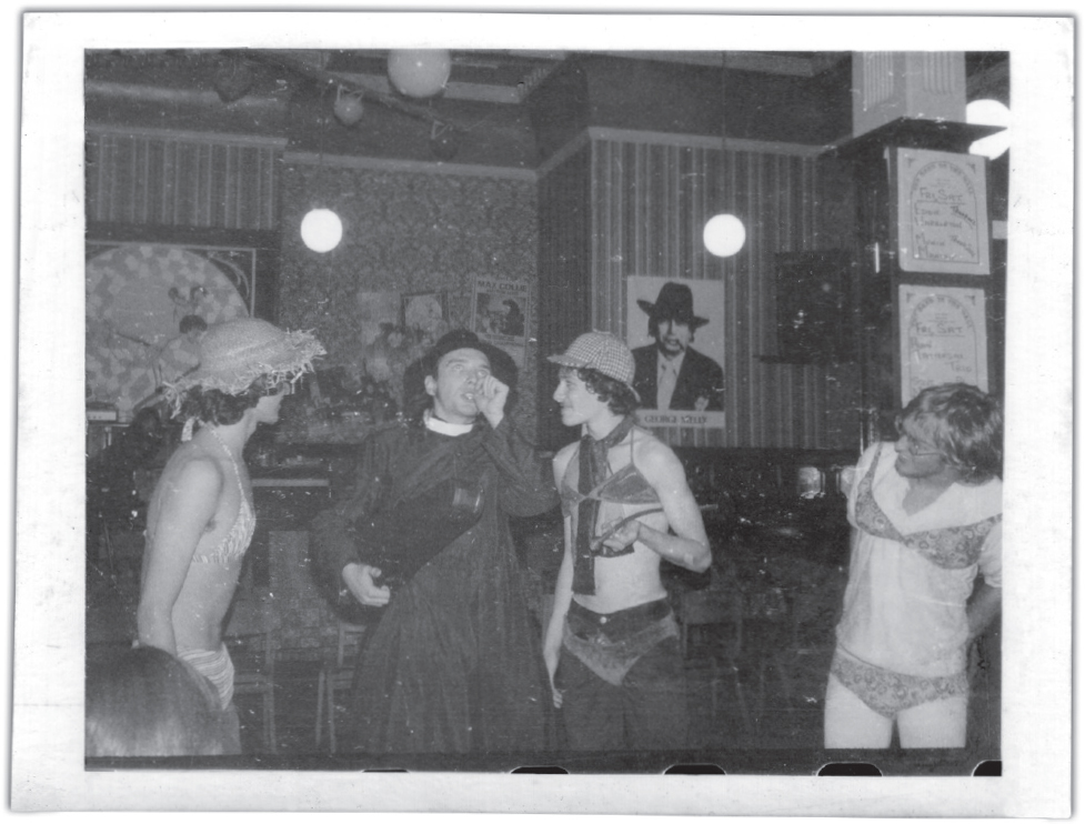 Start of image description, Twentieth Century Coyote perform at The Band on the Wall in Manchester. Rik Mayall stands centre-stage wearing a cassock and holding a violin case. The remaining members of the group wear bikinis over their clothing and a selection of hats including a deerstalker., end of image description