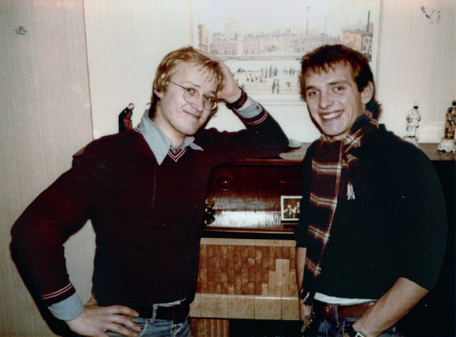 Start of image description, Adrian smiles and leans on the mantelpiece above the fireplace of Rik’s parents’ home in 1979. Mayall grins and wears a black and red striped scarf tight around his throat. A print of R S Lowry’s Northern River Scene hangs above them., end of image description