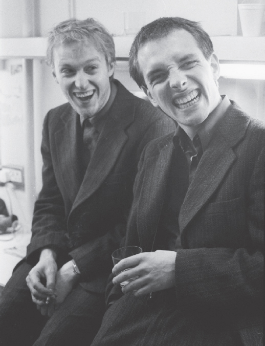 Start of image description, Rik grins maniacally and pulls a face and Adrian laughs as they sit backstage in their dressing room at the Comic Strip Club. The pair are wearing matching dark suits., end of image description