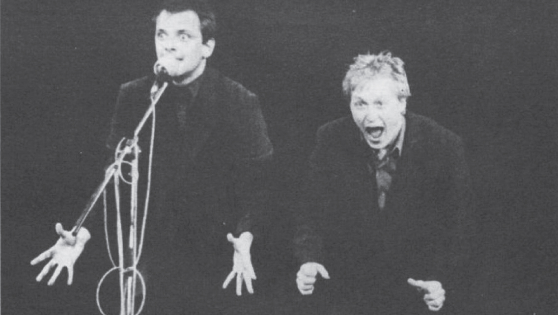 Start of image description, Adrian and Rik perform their Dangerous Brothers routine on stage. Adrian is leaning forward and screaming and Rik stands at the microphone, eyes wide and his fingers splayed at his sides., end of image description
