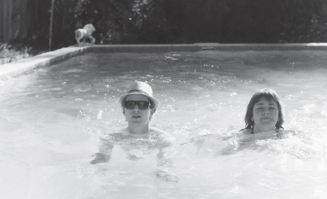 Start of image description, Adrian and fellow Comic Strip star, Dawn French, float in a swimming pool during a tour of Australia in 1981. Adrian wears a pair of sunglasses and a sun hat., end of image description