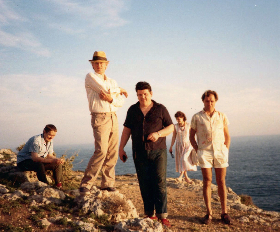 Start of image description, Adrian, arms folded and wearing a sun hat, stands on a rock at the edge of a cliff in Devon. Pete Richens is seated on a rock to his right, looking down. Robbie Coltrane is in the centre of the photo, with Pete Richardson on his left and Sophie Richardson visible in the background between them. The men are all in shirts and long trousers, except for Pete Richardson, who is wearing shorts that stop mid-thigh., end of image description