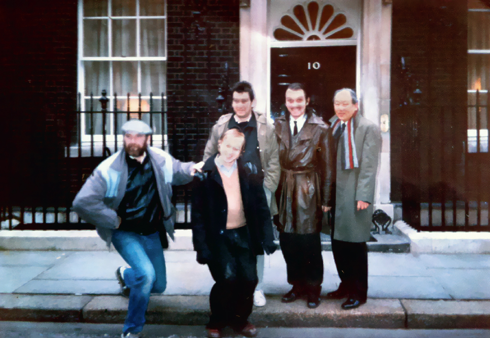 Start of image description, The group of 5 actors pose in front of the door to 10 Downing Street. Rik Mayall is wearing a knee-length, brown leather coat and is pulling a face at the camera., end of image description