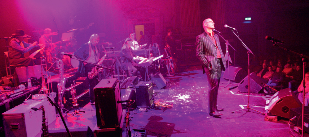 Start of image description, Adrian, wearing a dark pin-striped suit and red shirt, stands at the front of the stage and speaks into the microphone during a performance by the Bonzo Dog Doo-Dah Band at the London Astoria in 2006. The stage is bathed in pink light. The photograph is taken from the stage wings, with the rest of the band and their stage set-up on the left of the photo, and the audience visible on the right., end of image description