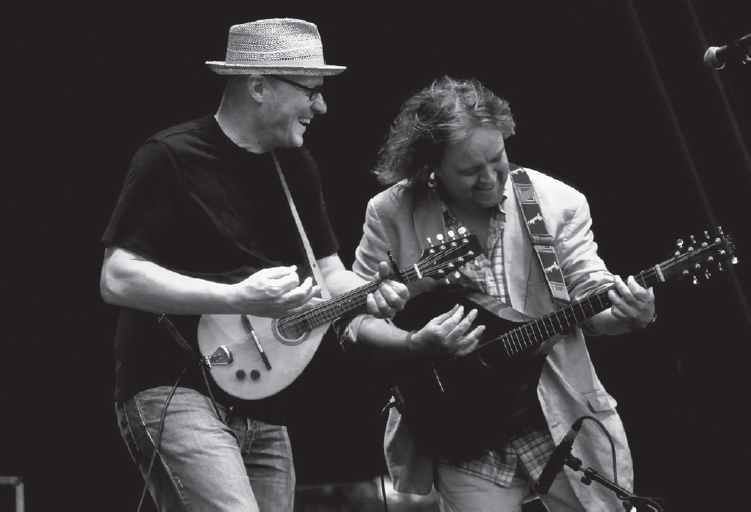 Start of image description, Adrian and Troy Donockley smile and laugh as they perform on stage as The Bad Shepherds. Adrian is shown playing a mandolin and Troy plays a cittern., end of image description