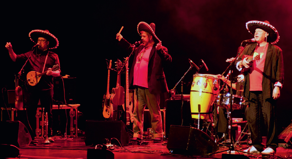 Start of image description, Adrian performs on stage with The Idiot Bastards. Adrian, Phill Jupitus and Neil Innes are wearing sombrero hats. Adrian and Neil both have mandolins, and Phill appears to be holding some sort of pipe. Behind Neil, Rowland Rivron is sat at a drum kit. Only his right arm and left hand are visible., end of image description