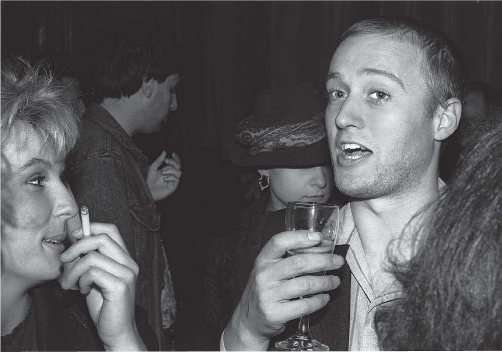 Start of image description, A younger-looking Adrian holds a glass of wine and is captured mid-sentence at a party. He isn’t wearing his glasses. Jennifer Saunders stands to the left, looking at Adrian and smiling as she smokes a cigarette. Other guests are visible behind them., end of image description
