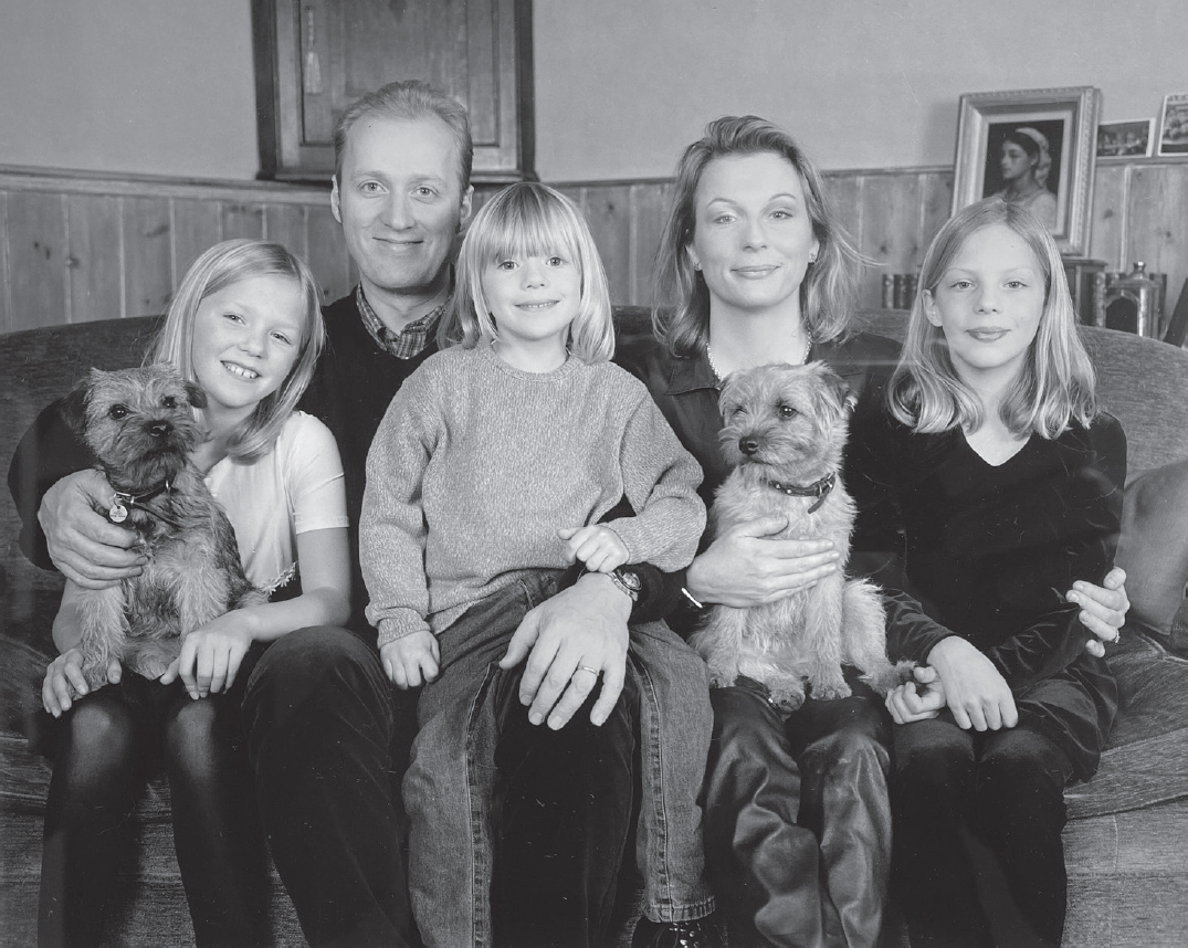 Start of image description, Adrian and Jennifer sit on the sofa with their 3 young daughters, Ella, Beattie, and Freya, and 2 small terriers, in the living room of their home. All three girls have shoulder-length blonde hair., end of image description