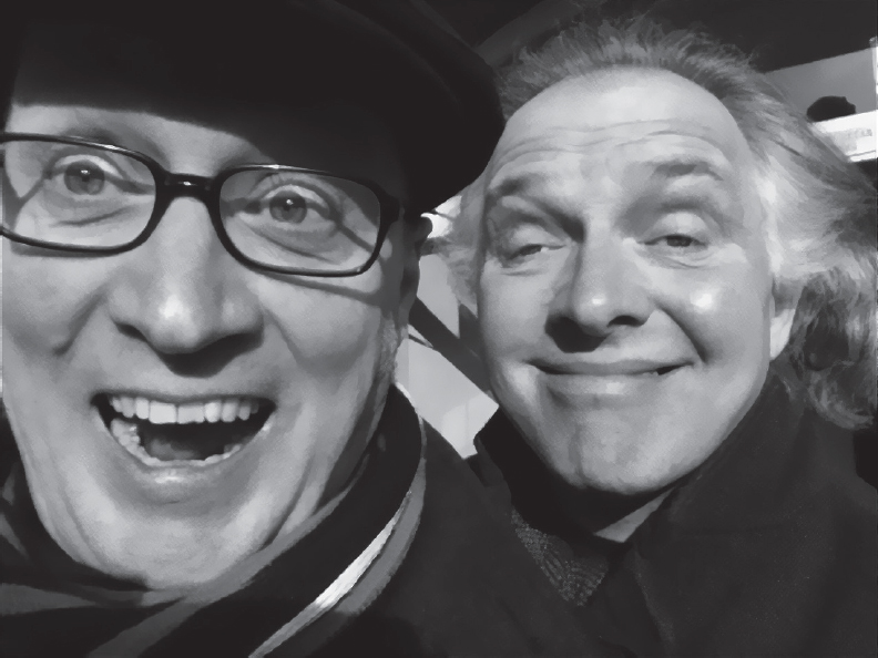 Start of image description, Adrian, wearing glasses and a flat cap, smiles with wide-open eyes and mouth as he takes a selfie of himself with Rik looking over his shoulder. Rik is giving a smarmy smile with his eyes half closed., end of image description
