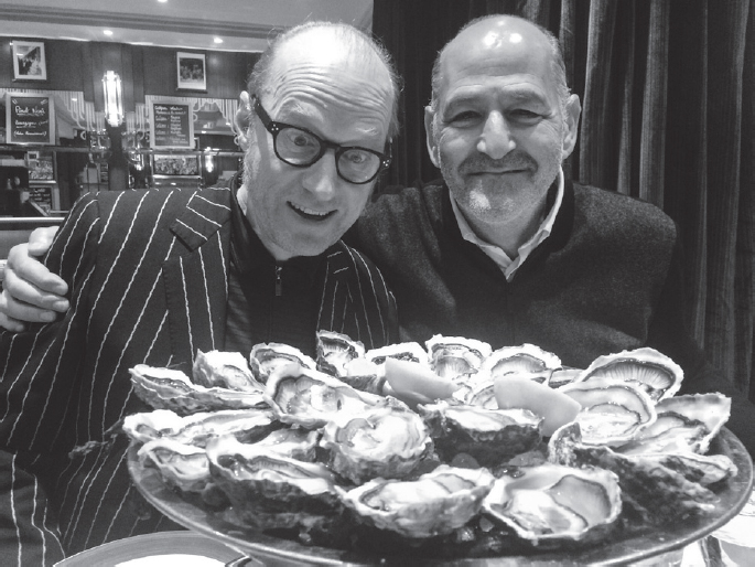 Start of image description, Adrian poses with his friend, David, and a huge platter of oysters in a restaurant in Paris. Adrian is wearing a pin-striped jacket and looking down at the oysters with an expression of amazement while David, in a jumper and shirt, smiles for the camera, end of image description